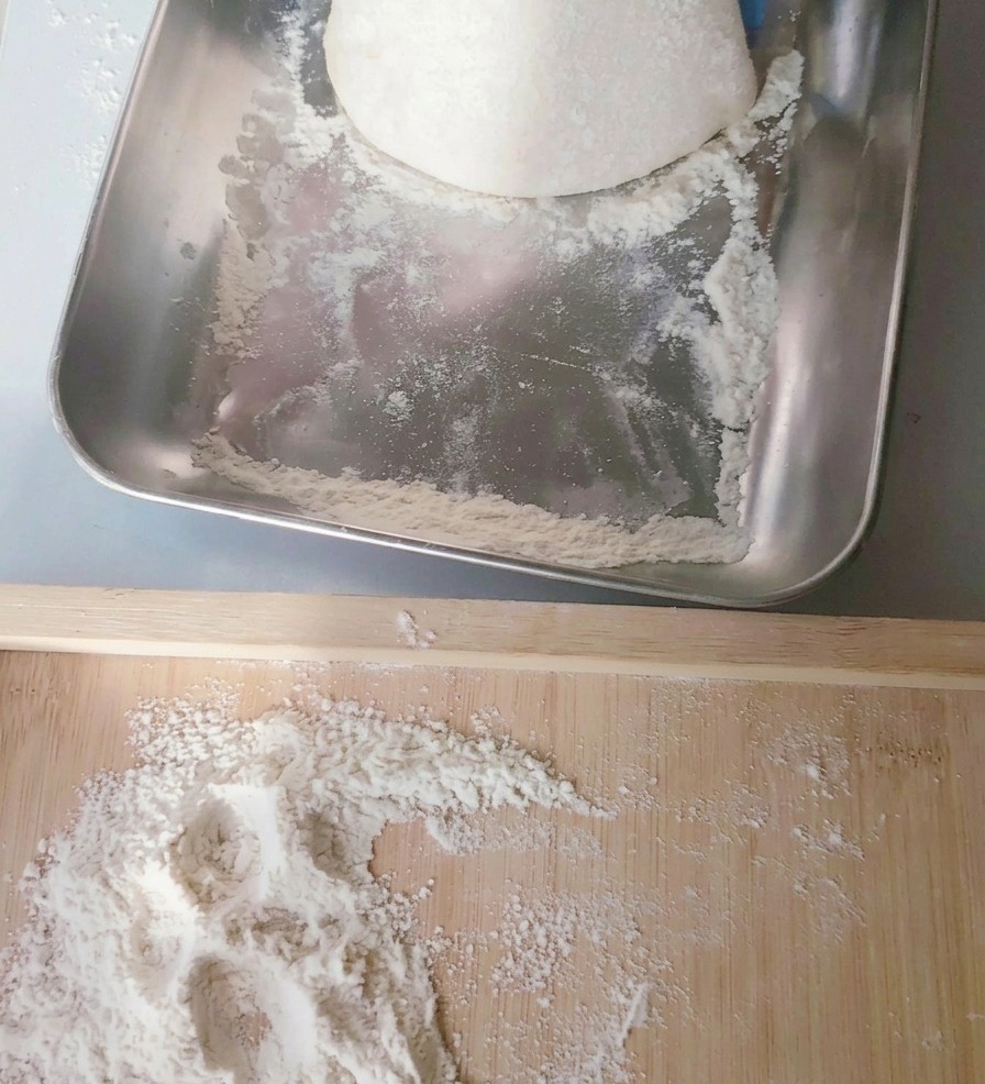 Eine runde Kugel Pizzateig wird aus einer Edelstahlwanne auf ein bemehltes Holzbrett gehoben.