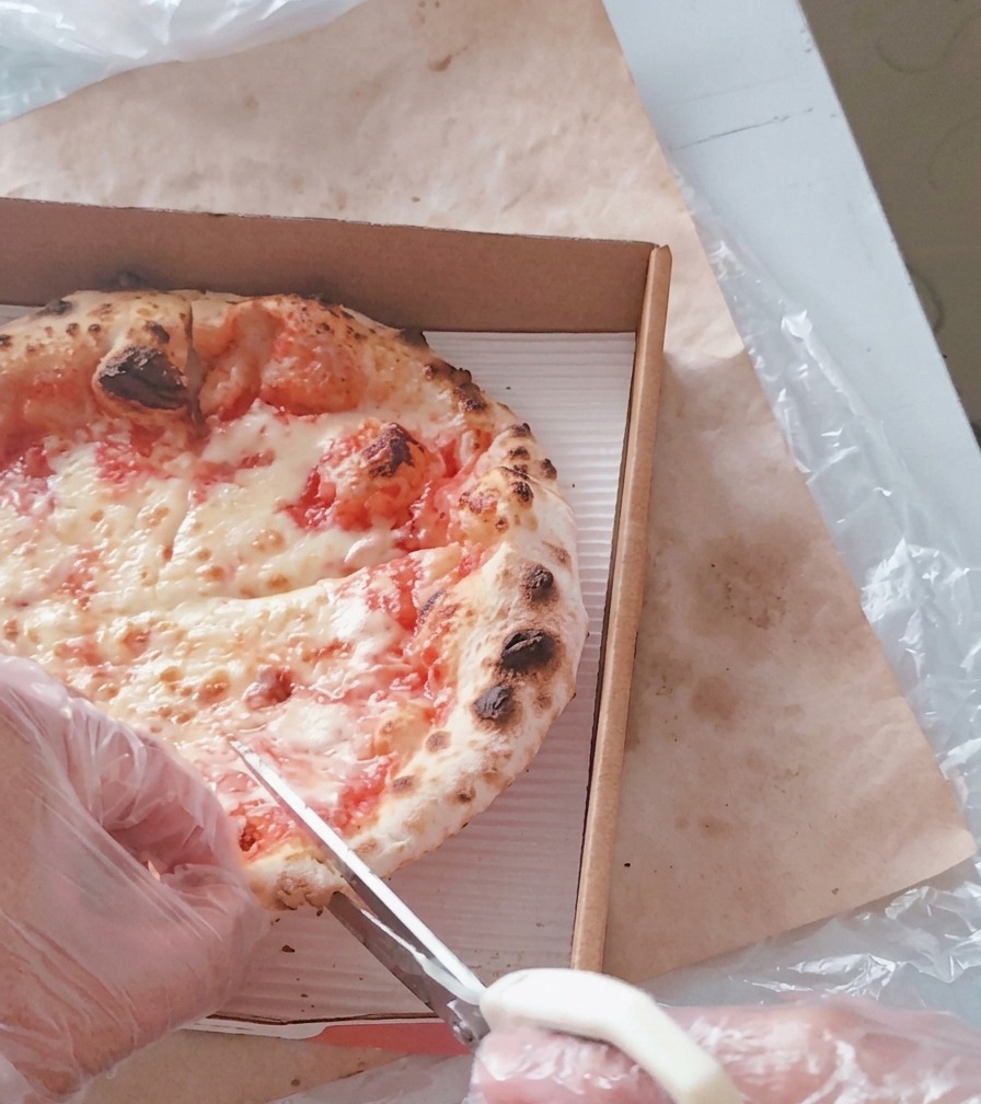 Eine person schneidet eine heiße Käsepizza mit einer weißen Küchenschere in einem Pizzakarton in Stücke.