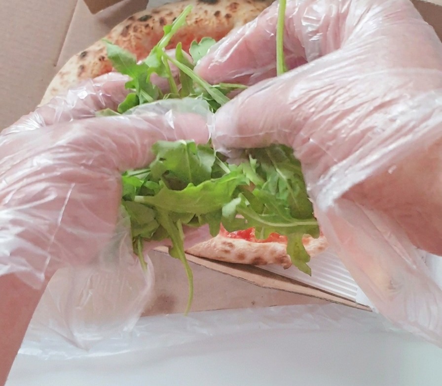 Hände in Plastikhandschuhen legen einen großen Haufen frische grüne Rucolablätter in die Mitte einer gebackenen Pizza im Karton.