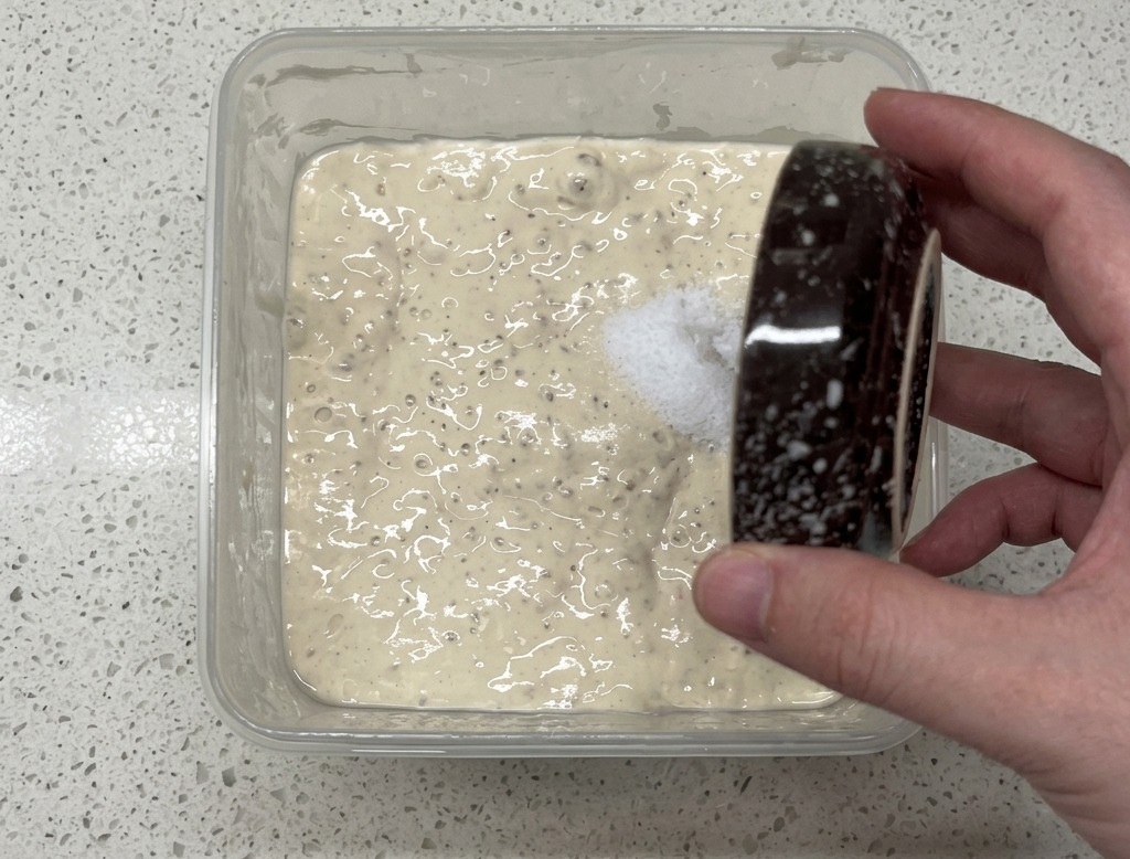 Eine Hand streut Salz aus einer kleinen Schüssel in einen Behälter mit blubberndem Poolish-Vorteig.