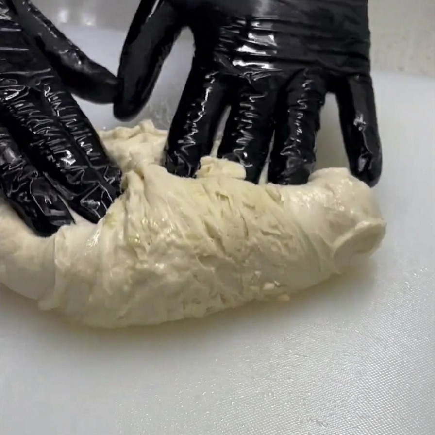 Eine Person mit schwarzen Handschuhen zieht und faltet ein Stück feuchten Teig auf einer weißen Oberfläche.