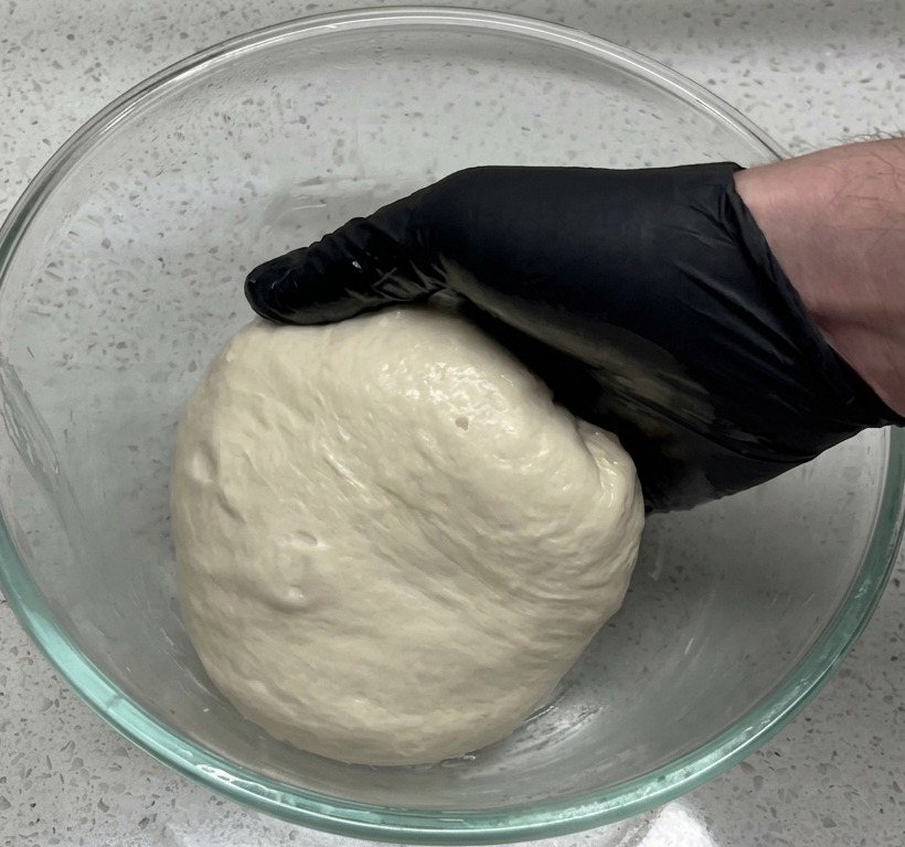 Eine Hand im schwarzen Handschuh legt eine glatte Teigkugel in eine große Glasschüssel auf einer Arbeitsplatte in Granitoptik.