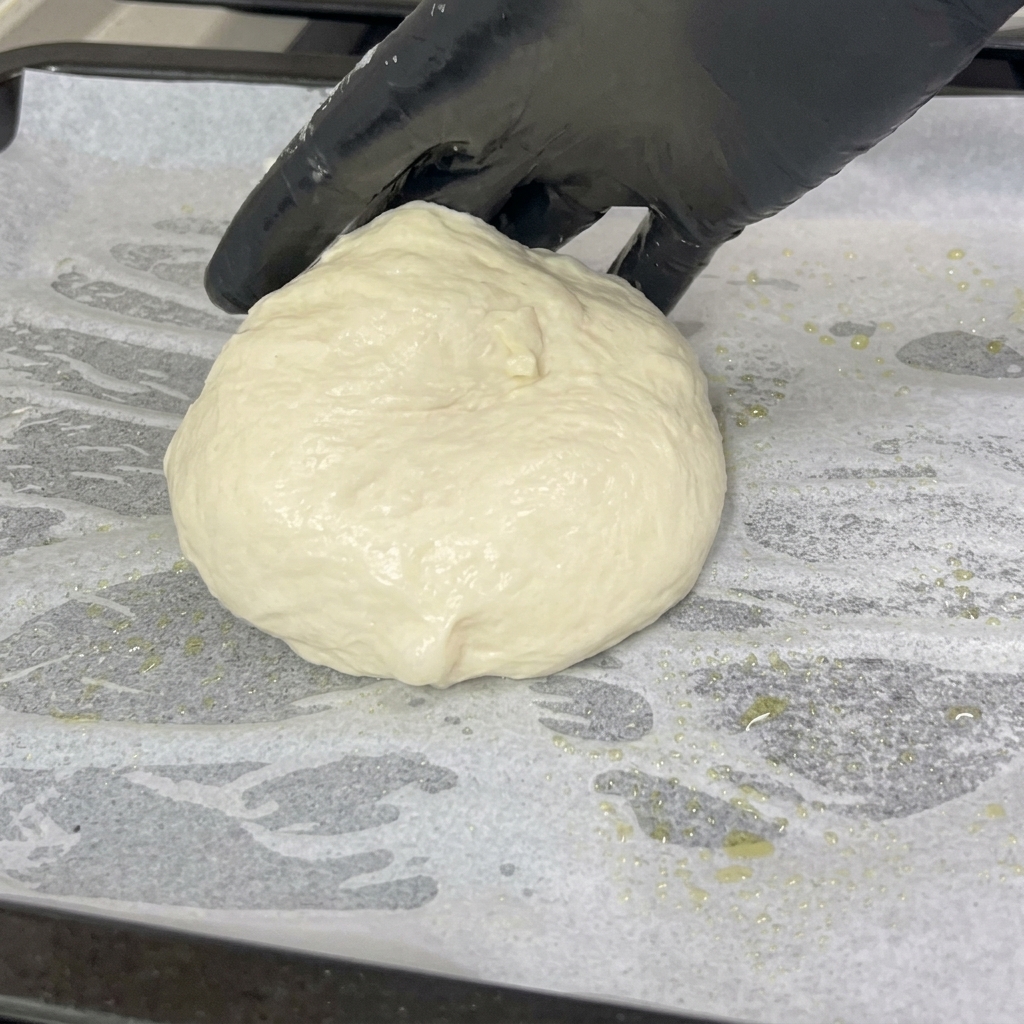 Eine behandschuhte Hand legt eine weiche, runde Pizzateigkugel auf ein mit Backpapier ausgelegtes Blech.