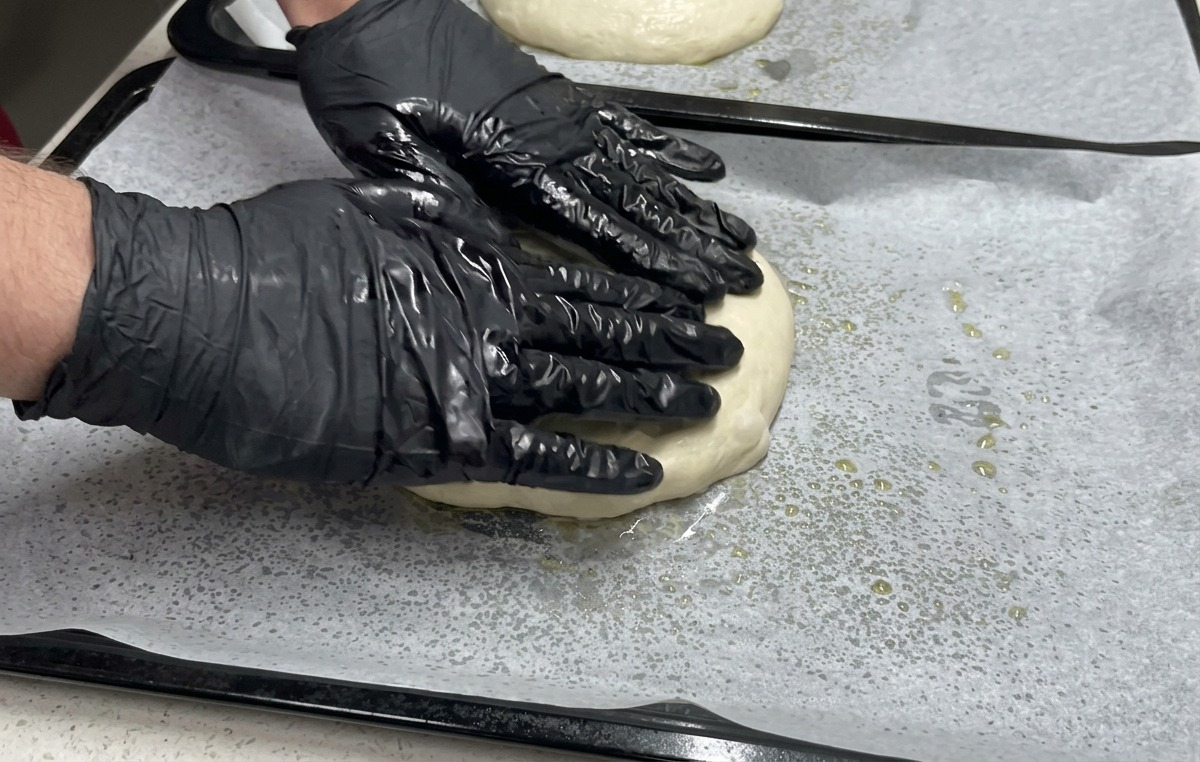 Zwei Hände in schwarzen Handschuhen drücken ein flaches Stück Teig auf einem mit Backpapier ausgelegten Backblech bis an die Ränder.