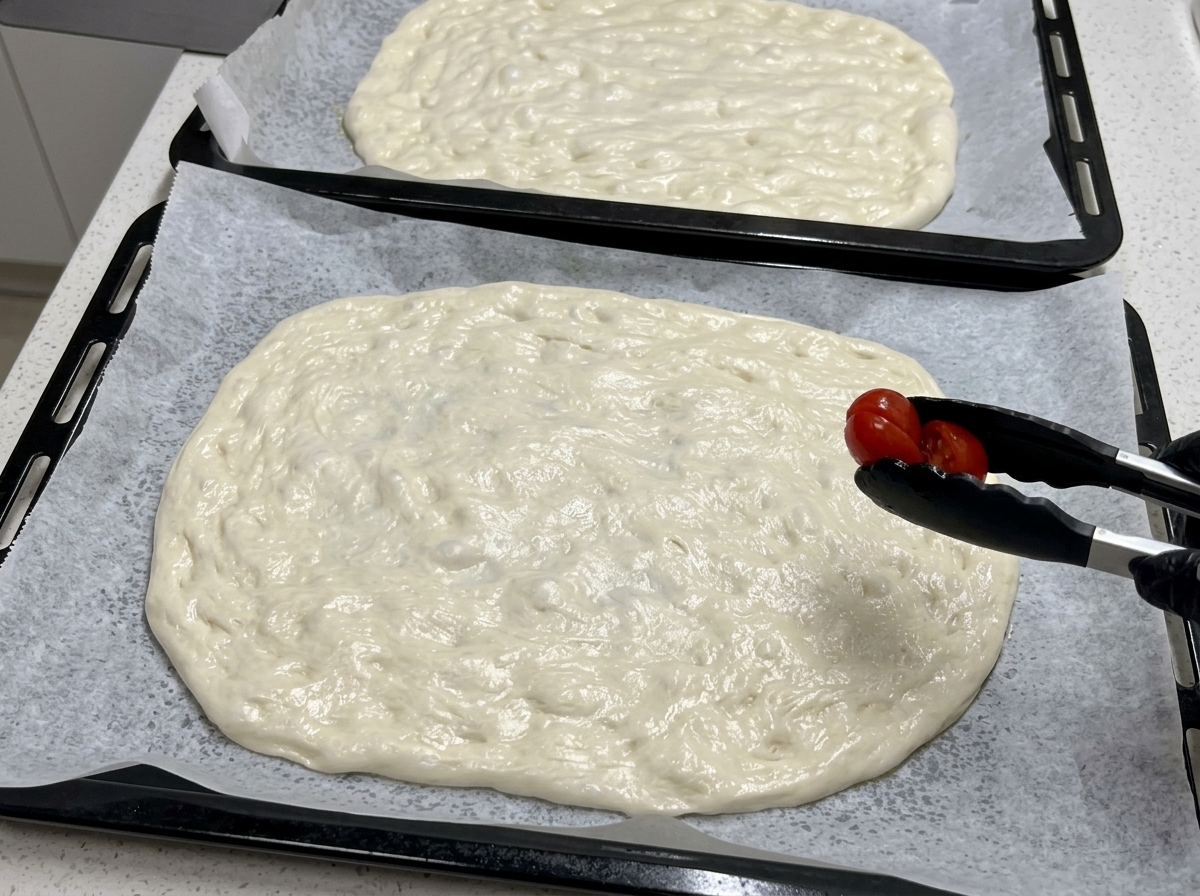 Mit einer schwarzen Küchenzange wird eine halbierte Kirschtomate auf einen großen, flachen, rohen Pizzateig gelegt.