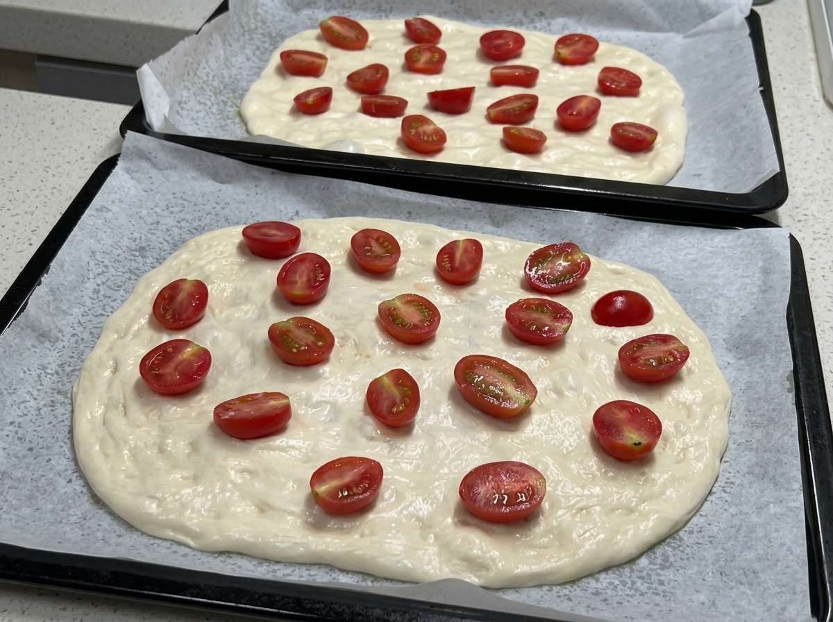 Zwei Backbleche zeigen flachgedrückten Pizzateig, belegt mit vielen halbierten Kirschtomaten, bereit zum Würzen.