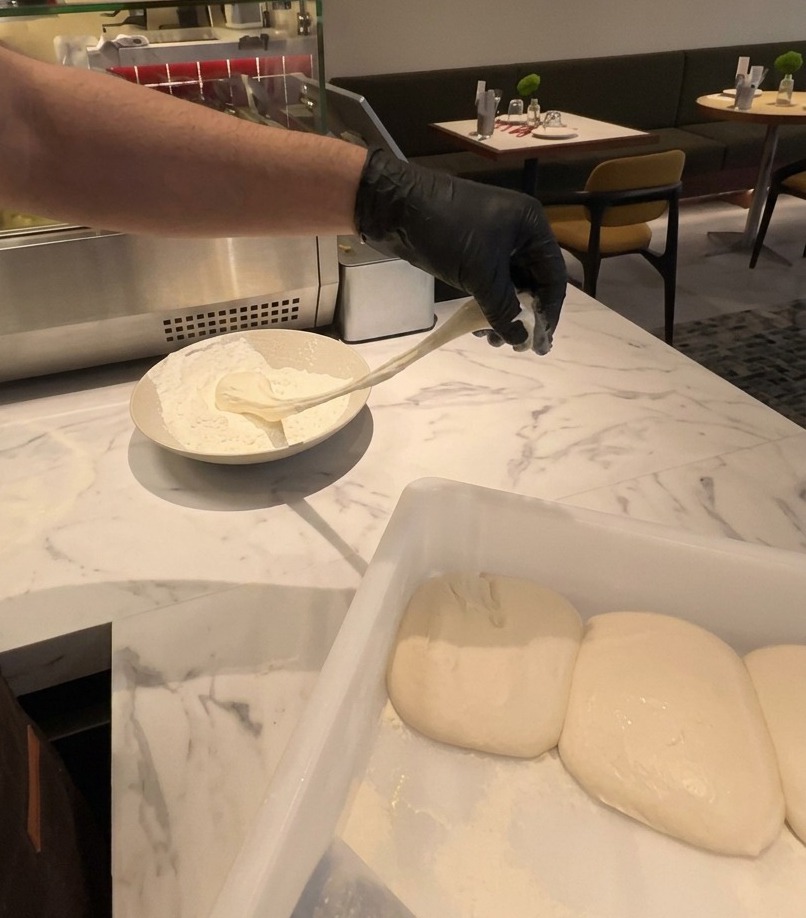 Eine Hand hebt ein dehnbares Stück fermentierten Pizzateig aus einer Schüssel mit weißem Mehl.