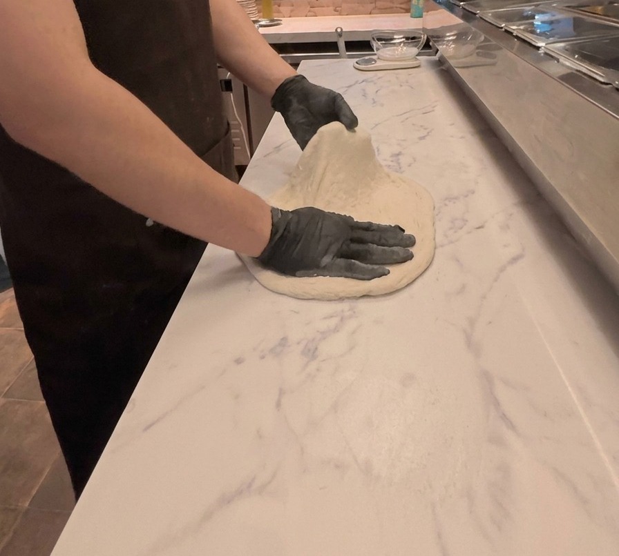 Ein Koch zieht und drückt Pizzateig mit beiden Händen auf einer weißen Arbeitsplatte mit Marmormuster flach.
