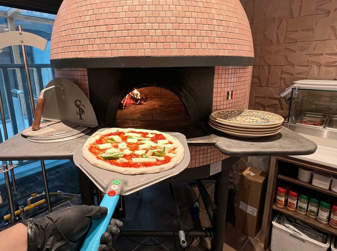 Eine ungebackene Pizza mit Tomatensoße und Mozzarella liegt auf einem Metallschieber am Eingang eines rosa gefliesten Holzofens.