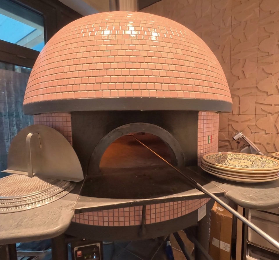 Das Äußere eines rosa gefliesten Kuppelofens für Pizzen, in dem eine Pizza in der Nähe der glühenden Kohlen backt.