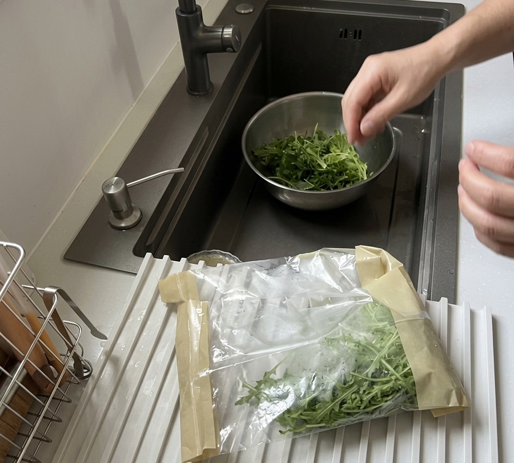 Frischer Rucola wird in einer Edelstahlschüssel unter fließendem Leitungswasser gewaschen.
