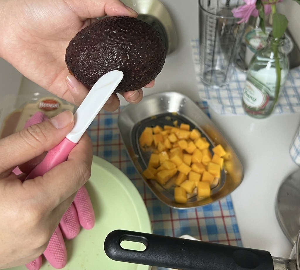 Hände schneiden eine geschälte Avocadohälfte über einer Schale mit gewürfelter Mango in kleine Stücke.