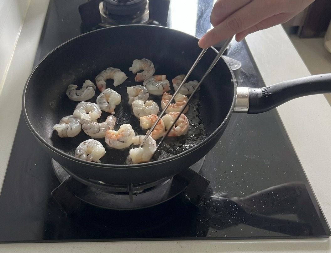 Frische Garnelen werden in einer dunklen Pfanne mit Olivenöl angebraten, einige färben sich bereits rosa und werden undurchsichtig.