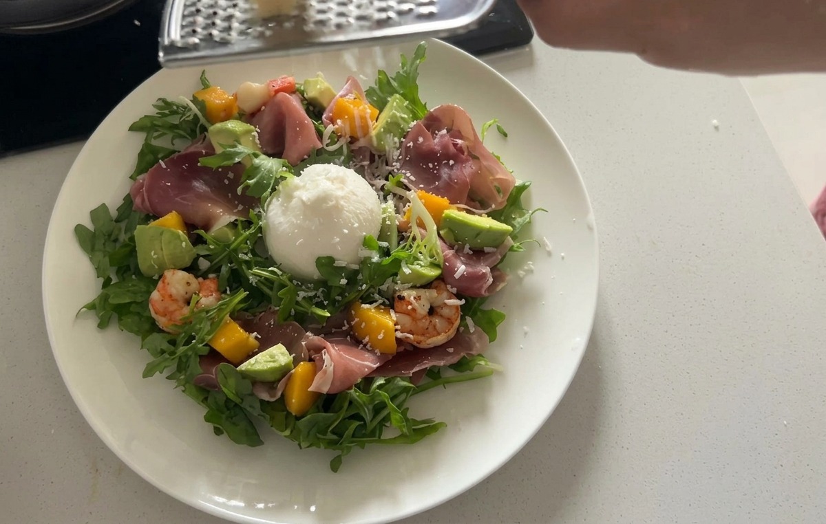 Ein angerichteter Rucolasalat, garniert mit einer großen weißen Burrata-Kugel und feinen Streifen von luftgetrocknetem Schinken.