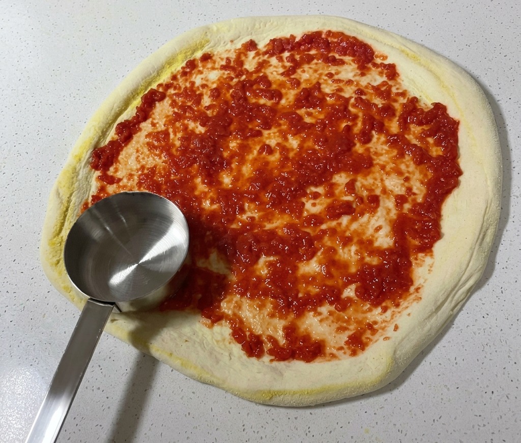 Eine Schöpfkelle verteilt rote Tomatensauce kreisförmig auf dem rohen Pizzateig.