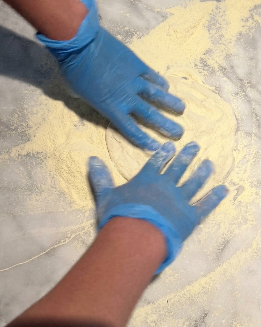Hände mit blauen Handschuhen drücken eine runde Pizzateigkugel in eine dicke Schicht Semolina-Mehl auf einer Marmorarbeitsplatte.