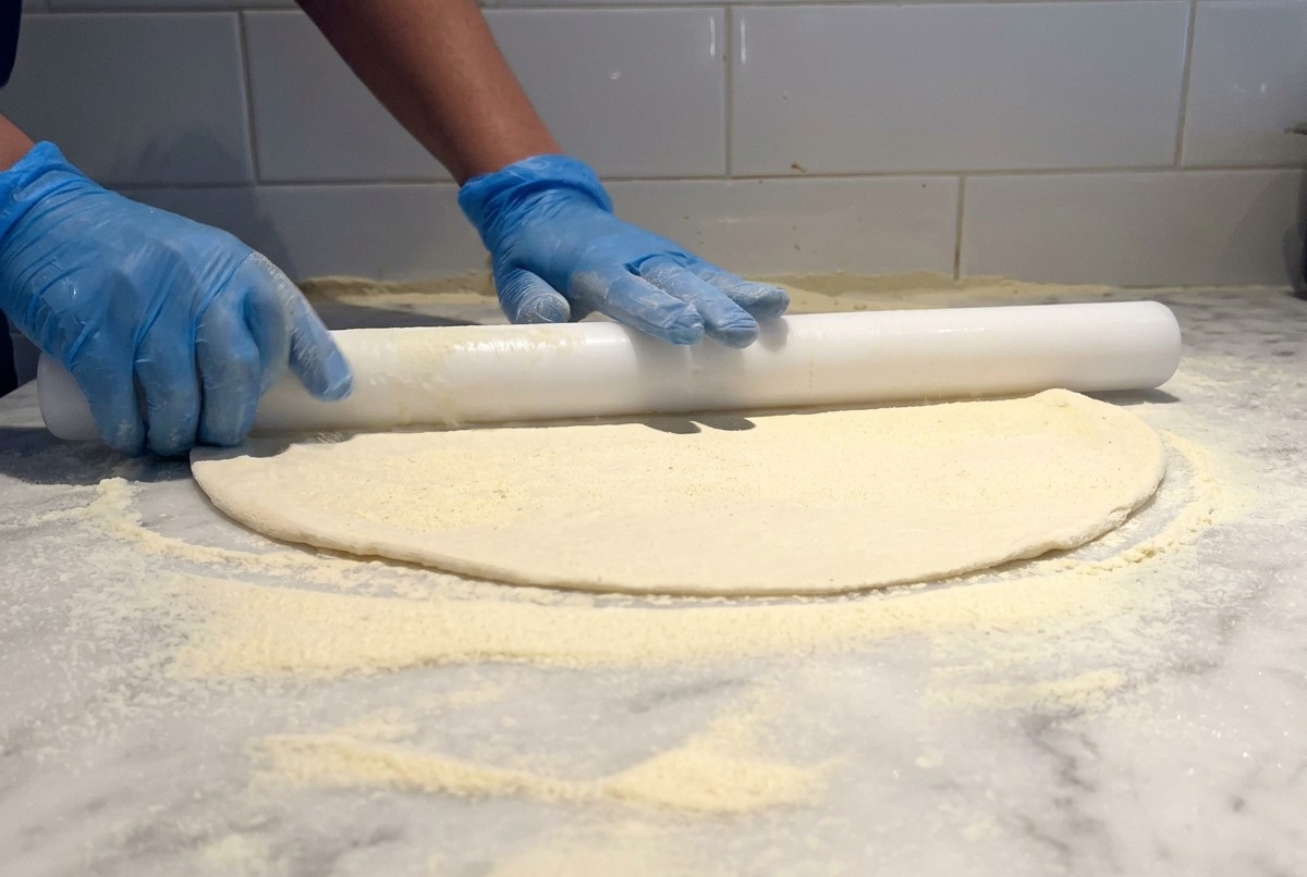 Ein weißes Nudelholz rollt einen dünnen Pizzateig auf einem bemehlten Marmortisch aus.