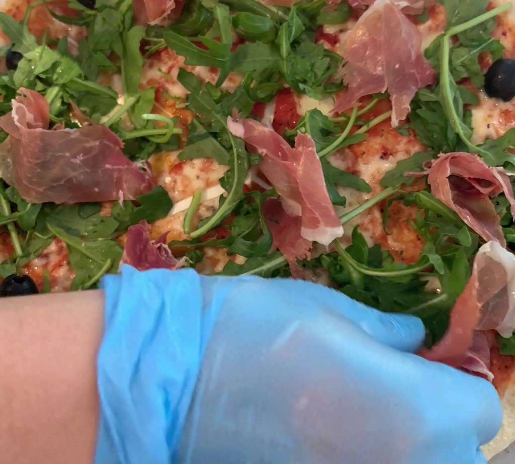Eine Hand in Handschuhen legt vorsichtig dünne Parmaschinkenscheiben auf die mit Rucola belegte Pizza.