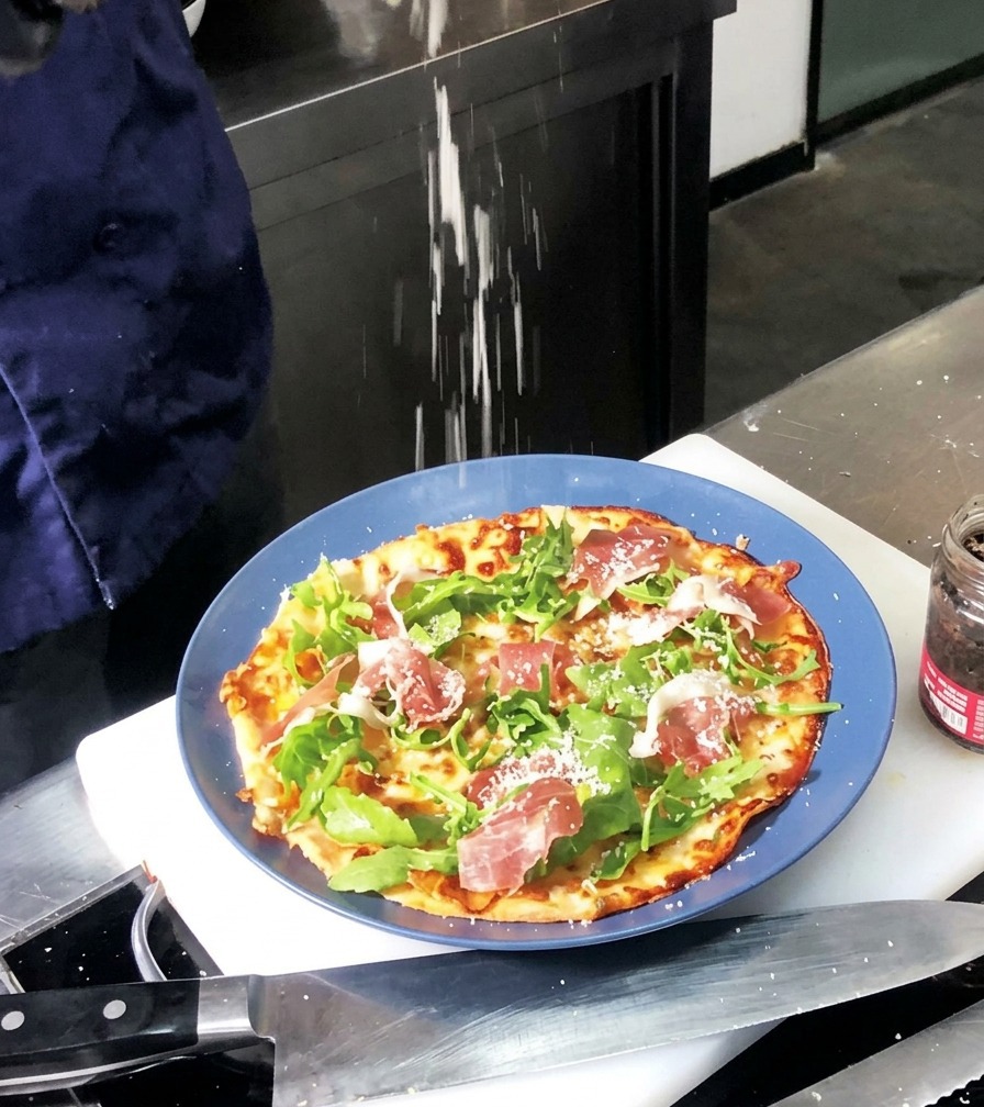 Feiner weißer Parmesan wird über eine gebackene Pizza gestreut, die mit grünem Rucola und roten Schinkenscheiben belegt ist.