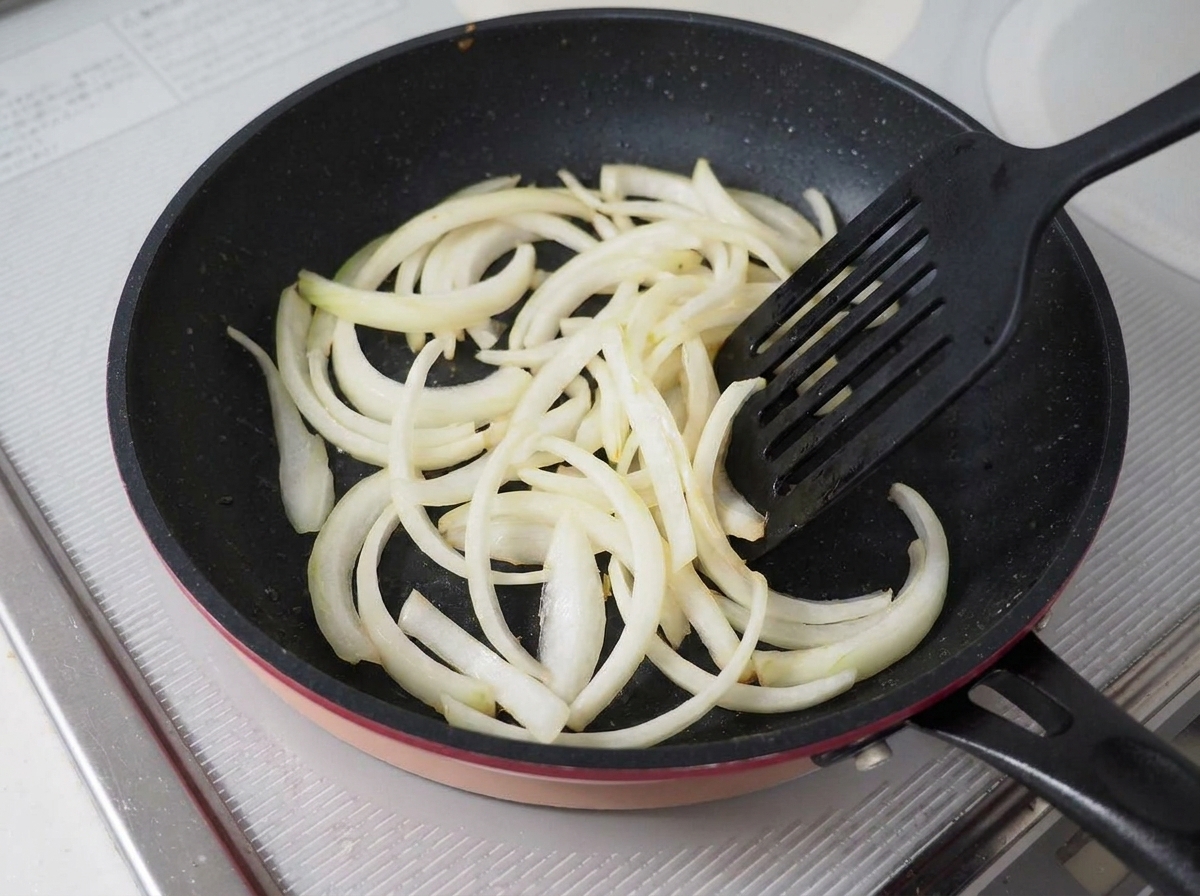 Geschnittene Zwiebeln werden in einer dunklen Antihaftpfanne mit einem schwarzen Pfannenwender angebraten.