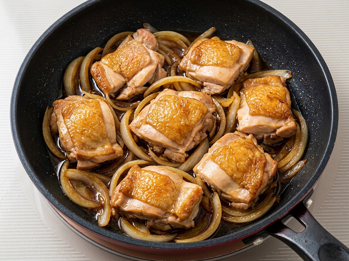 Gebratene Hähnchenstücke, die mit Zwiebeln und dunkler Sauce in einer Pfanne köcheln.