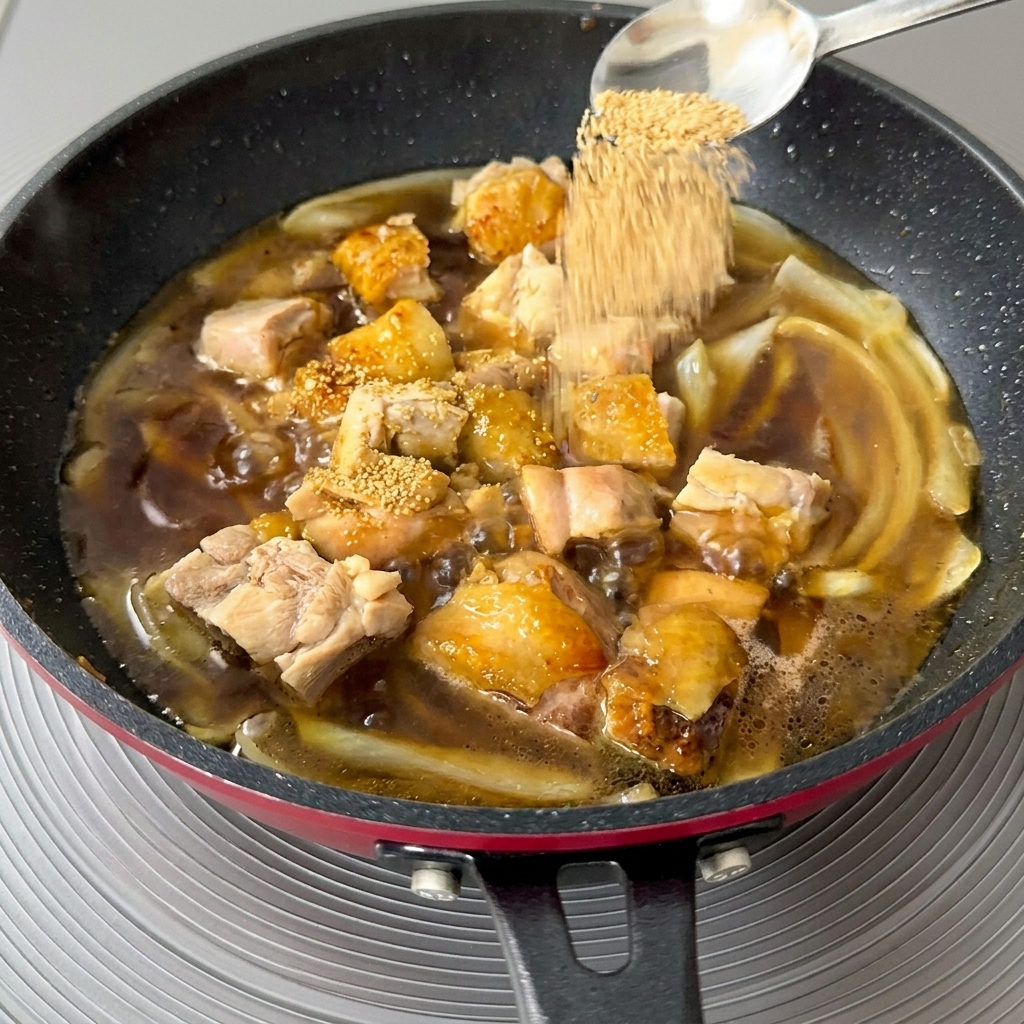 Nahaufnahme, wie ein Löffel mit goldenem Dashi-Granulat in eine köchelnde Pfanne mit Hähnchen und Zwiebeln gestreut wird.