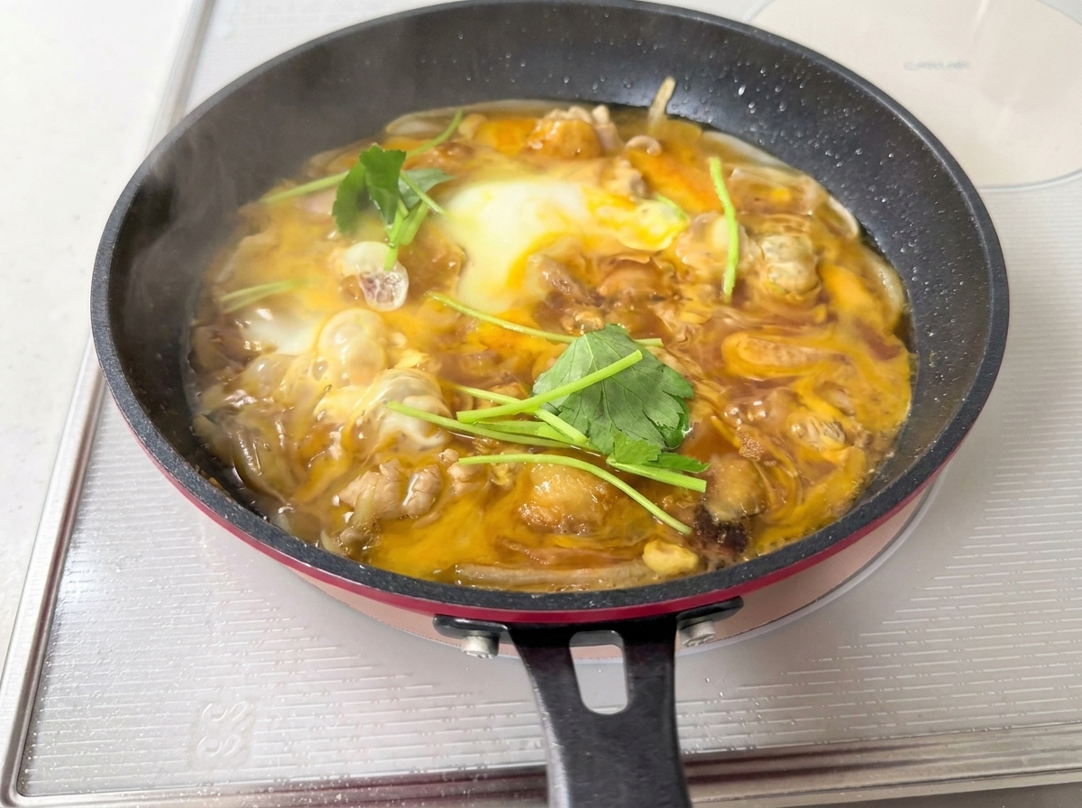 Frische grüne Mitsuba-Blätter werden über eine Pfanne mit weich gestocktem Ei und Hähnchen gestreut.
