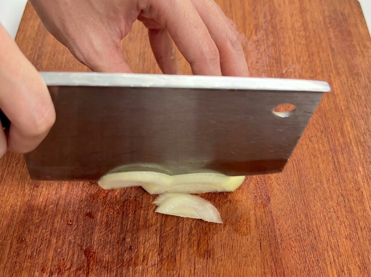 Nahaufnahme einer weißen Zwiebel, die auf einem Holzbrett in dünne Streifen geschnitten wird.