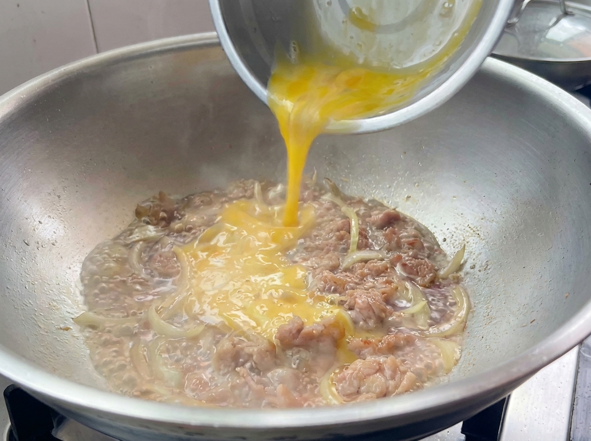 Verquirlte Eimischung wird über das schmorende Schweinefleisch mit Zwiebeln in der Pfanne geträufelt.