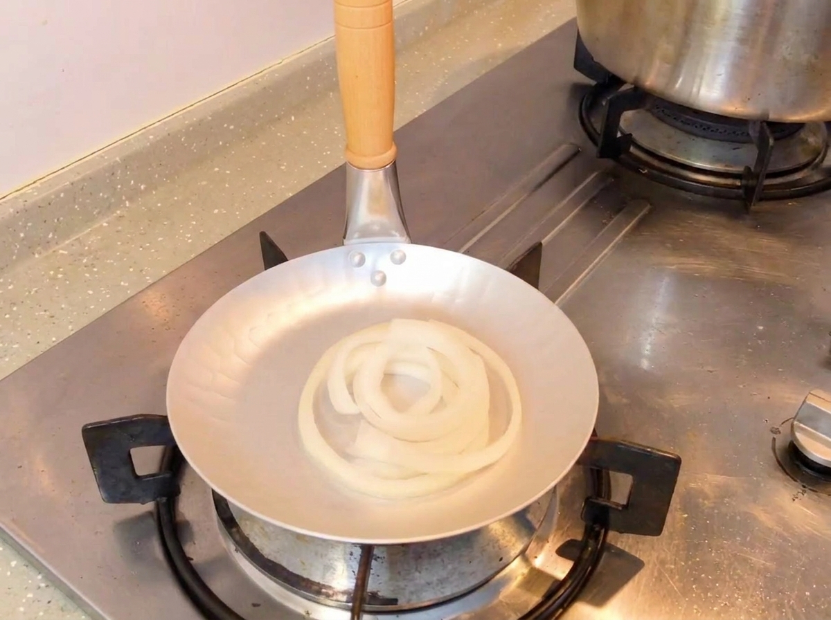 Die dunkle Sojasaucenmischung wird von einem Löffel in eine Pfanne mit rohen weißen Zwiebelringen gegossen.