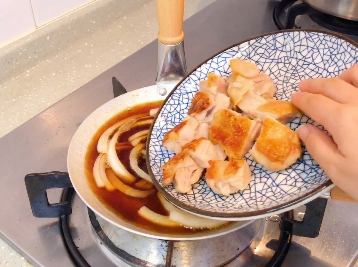 Knusprig gebratene Hähnchenstücke werden in eine Pfanne mit köchelnden Zwiebeln und einer Sojasauce gegeben.