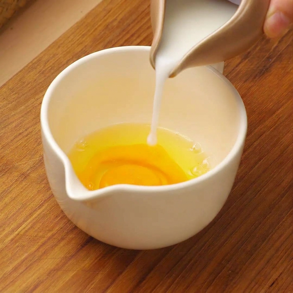 Weiße Milch wird aus einem kleinen Krug in einen weißen Messbecher mit aufgeschlagenen rohen Eiern gegossen.