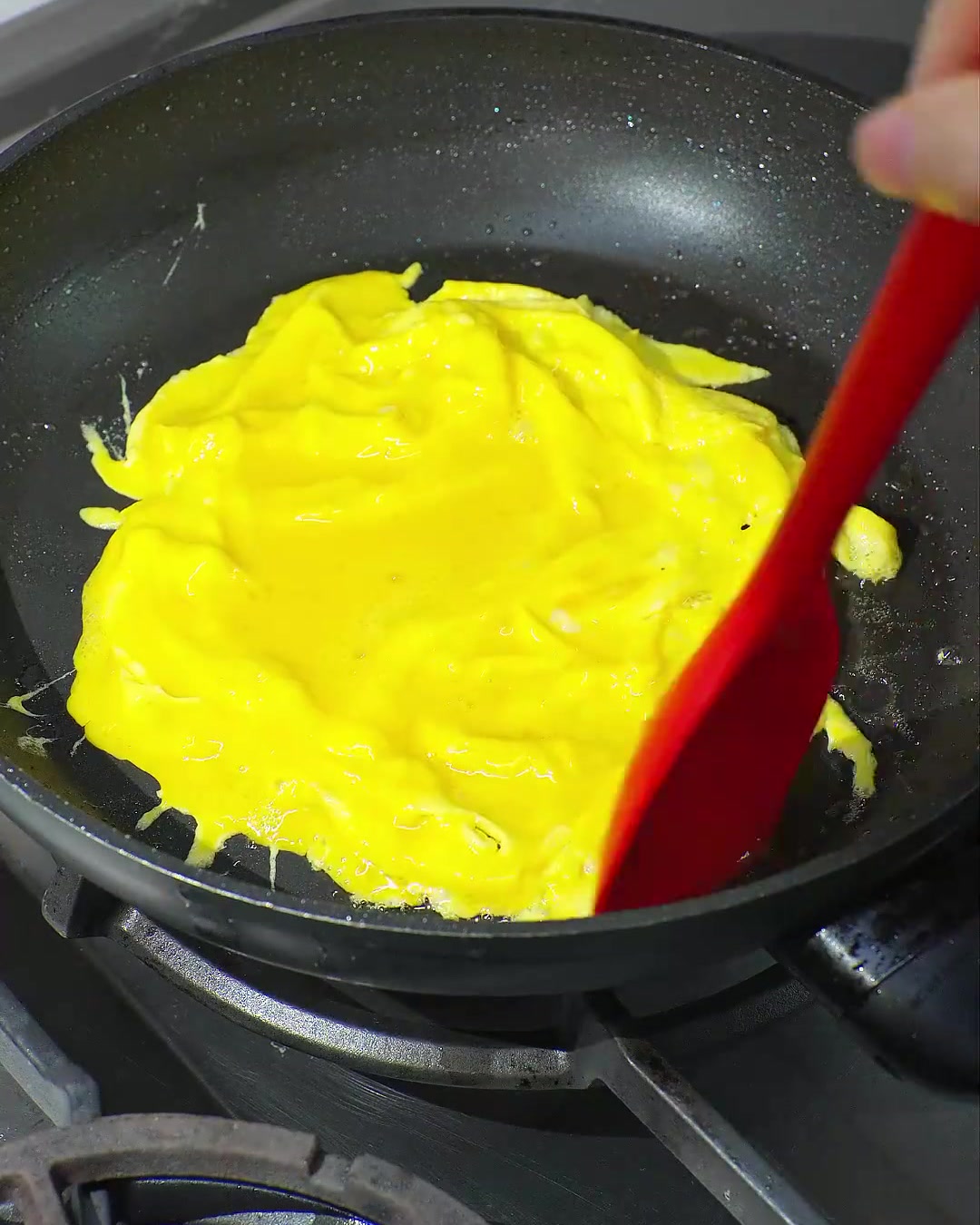 Ein roter Spatel faltet ein leuchtend gelbes, teilweise gekochtes Rührei in einer schwarzen Bratpfanne zusammen.