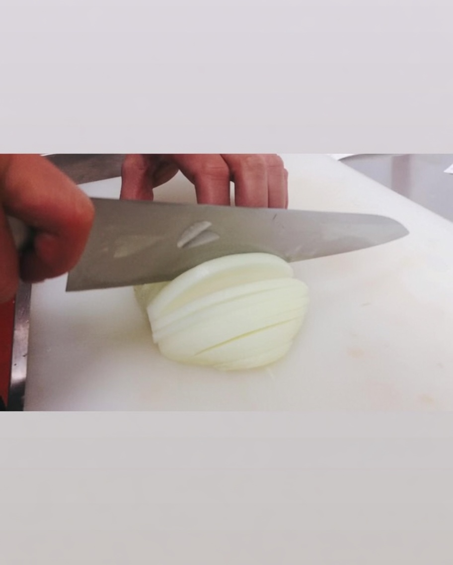 Eine rohe weiße Zwiebel wird mit einem großen Messer auf einem weißen Schneidebrett in Streifen geschnitten.