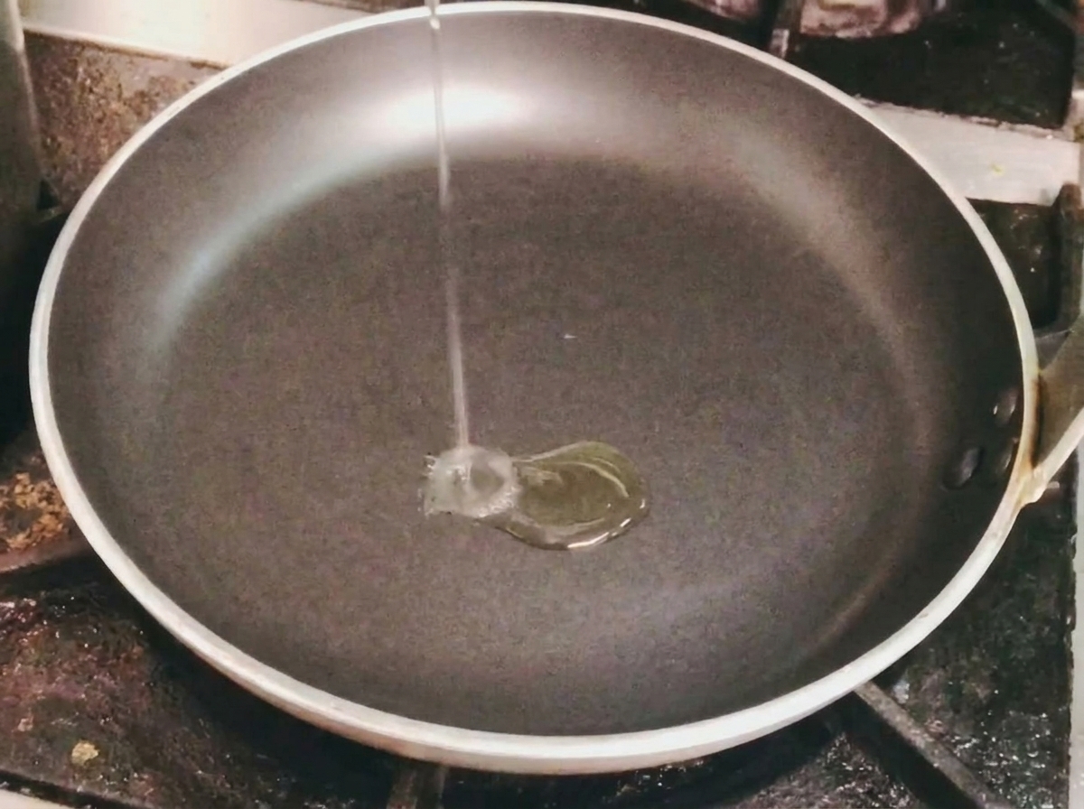 Ein dünner Strahl Speiseöl wird in eine leere schwarze Bratpfanne auf einem Herd gegossen.
