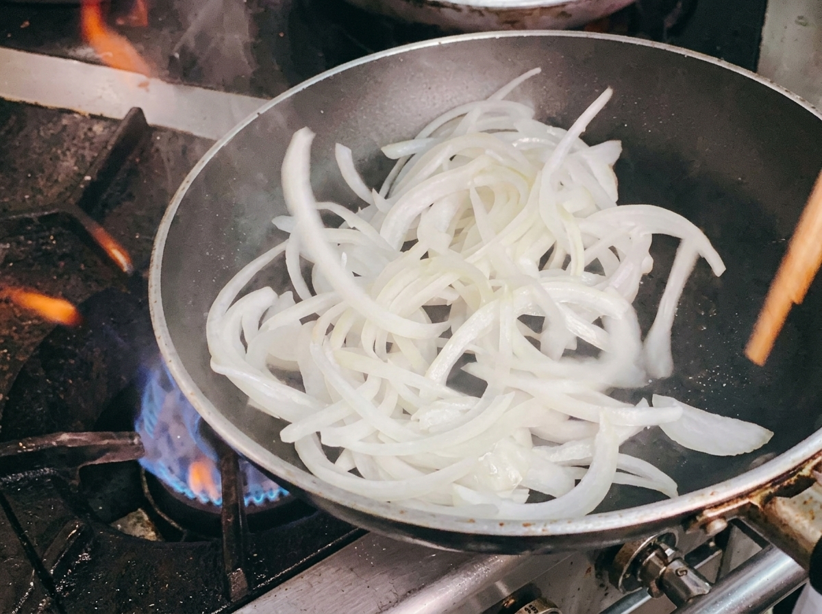 Die geschnittenen weißen Zwiebeln werden in der Pfanne mit Holzstäbchen angebraten.