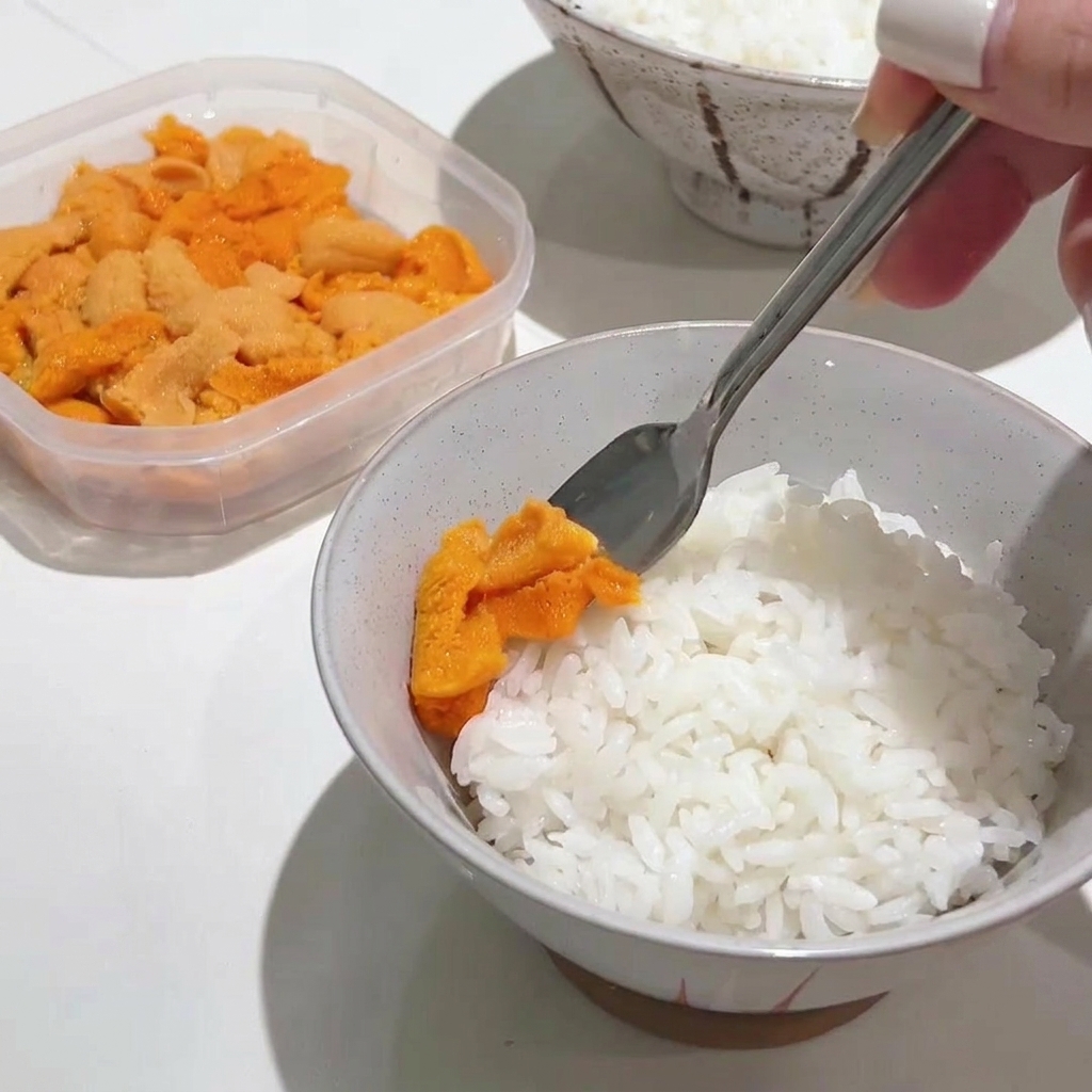 Ein Metalllöffel platziert behutsam eine großzügige Portion leuchtend orangefarbenen Seeigel auf einer Schüssel mit heißem weißem Reis.