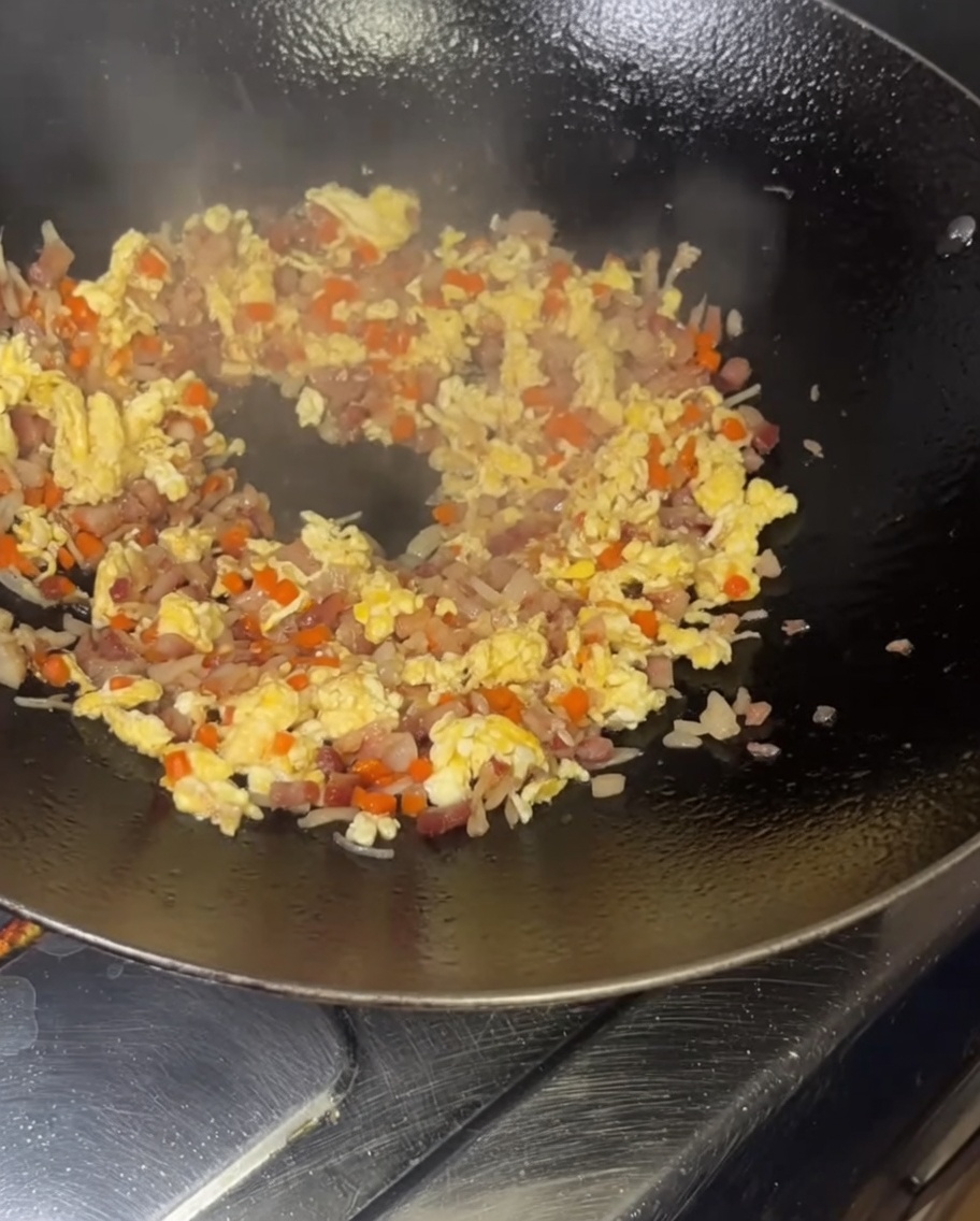 Eine Schüssel mit gelben, eiumhüllten Reiskörnern wird in einen rauchend heißen Wok gefüllt mit gebratenen Zutaten geleert.