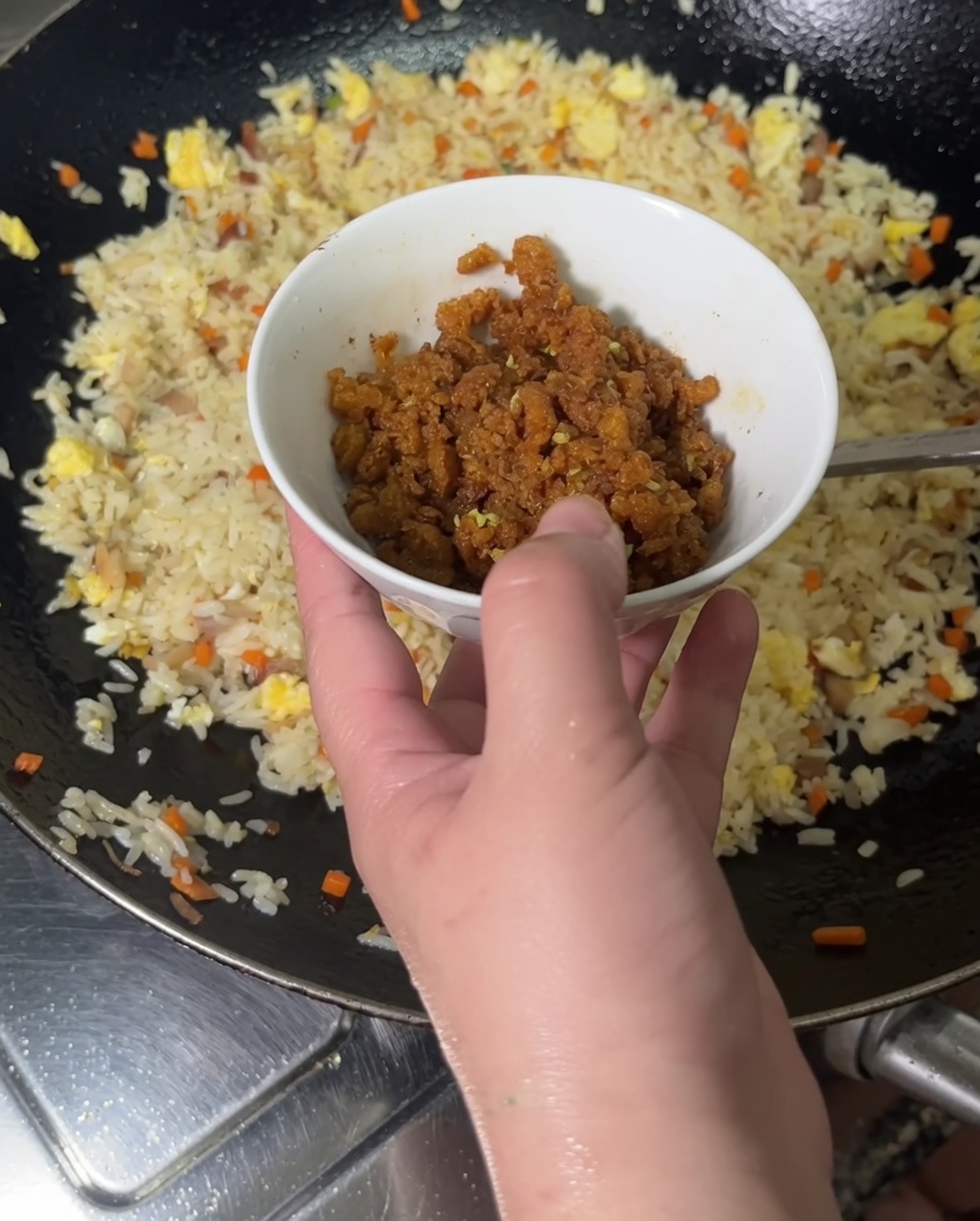 Eine Hand gießt gekochten, leuchtend orangefarbenen Seeigelrogen aus einer kleinen Schüssel in einen Wok mit gebratenem Reis.