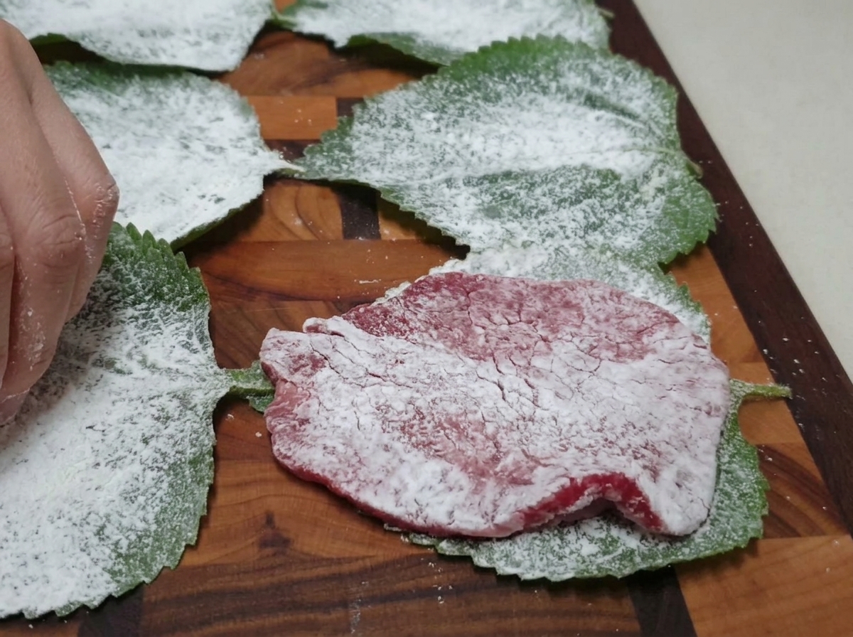 Eine bemehlte Rindfleischscheibe wird auf einem bemehlten grünen Perillablatt auf einem Holzschneidebrett platziert.