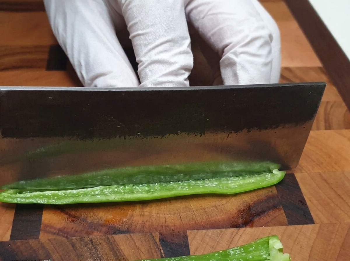 Ein Kochmesser schneidet eine grüne Chilischote fein auf einem Holzschneidebrett.
