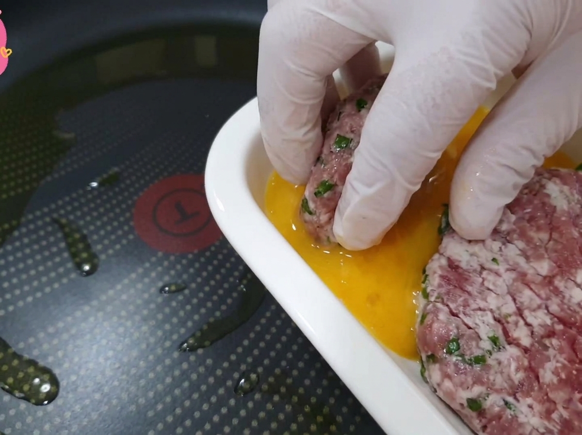 Eine Hand in einem transparenten Handschuh taucht ein Stück Rindfleisch mit Perillablatt in eine Schale mit goldener Eiermasse.