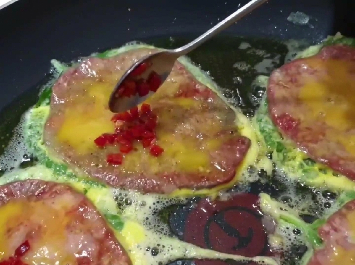 Nahaufnahme eines Löffels, der gehackte grüne und rote Chilis auf die in der Pfanne bratenden Rindfleisch-Pfannkuchen gibt.