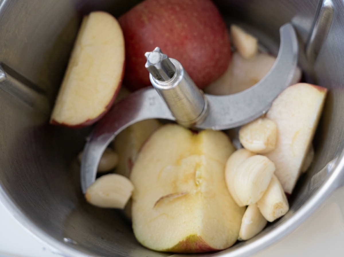 Apfelscheiben und geschälte Knoblauchzehen in der Schüssel einer Küchenmaschine vor dem Pürieren.