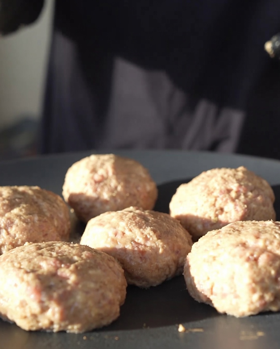 Runde Fleisch-Patties, die in einer flachen, schwarzen Gusseisenpfanne bei schwacher Hitze braten.