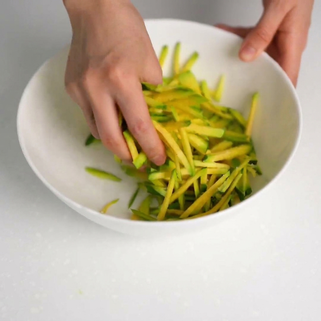 Hände drücken gesalzene Zucchinistreifen aus, um die Flüssigkeit in eine saubere Schüssel abzugießen.
