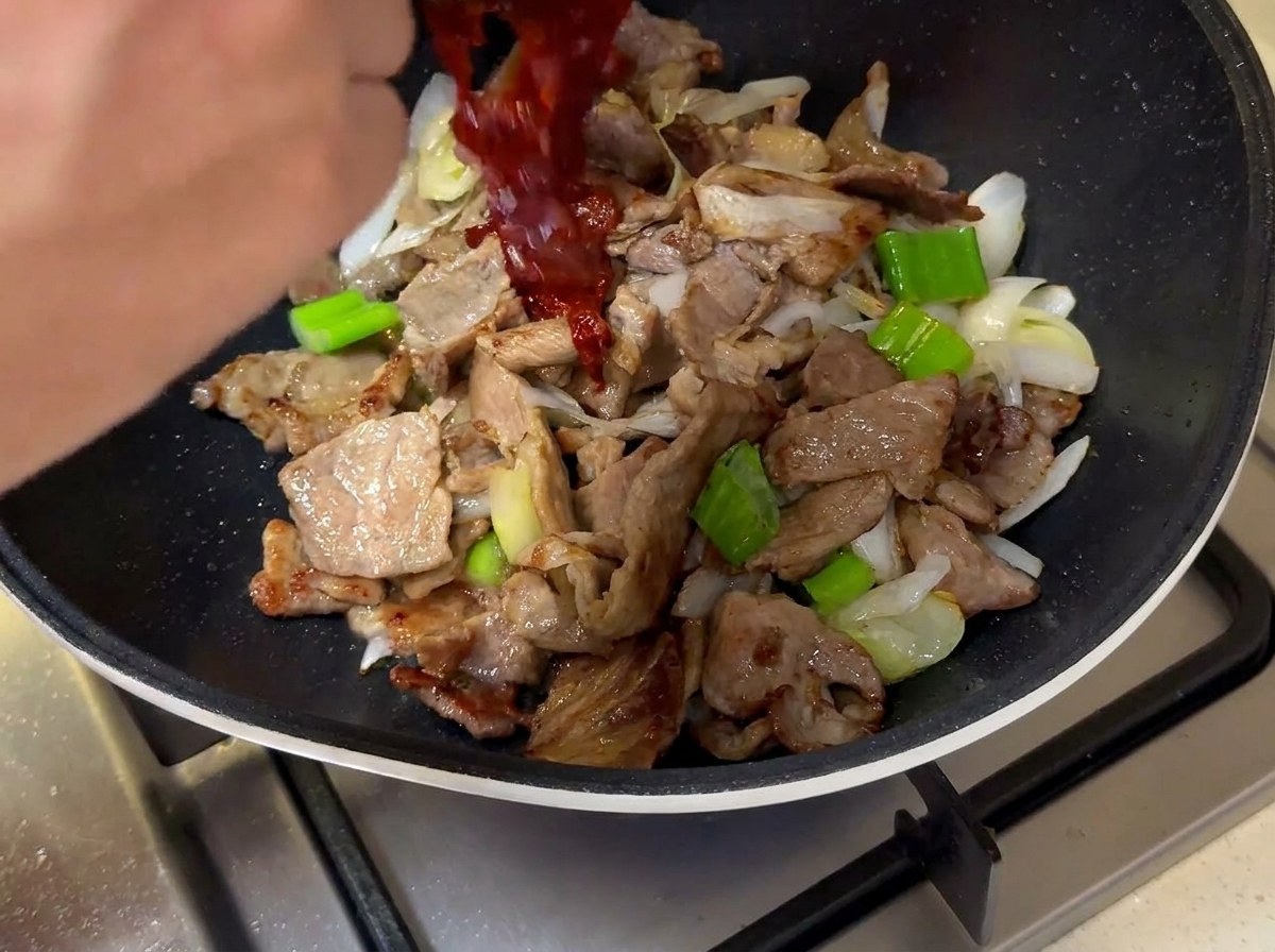 Die dunkelrote Sauce wird aus einer kleinen Schüssel über das Fleisch und die Zwiebeln in die Pfanne gegossen.
