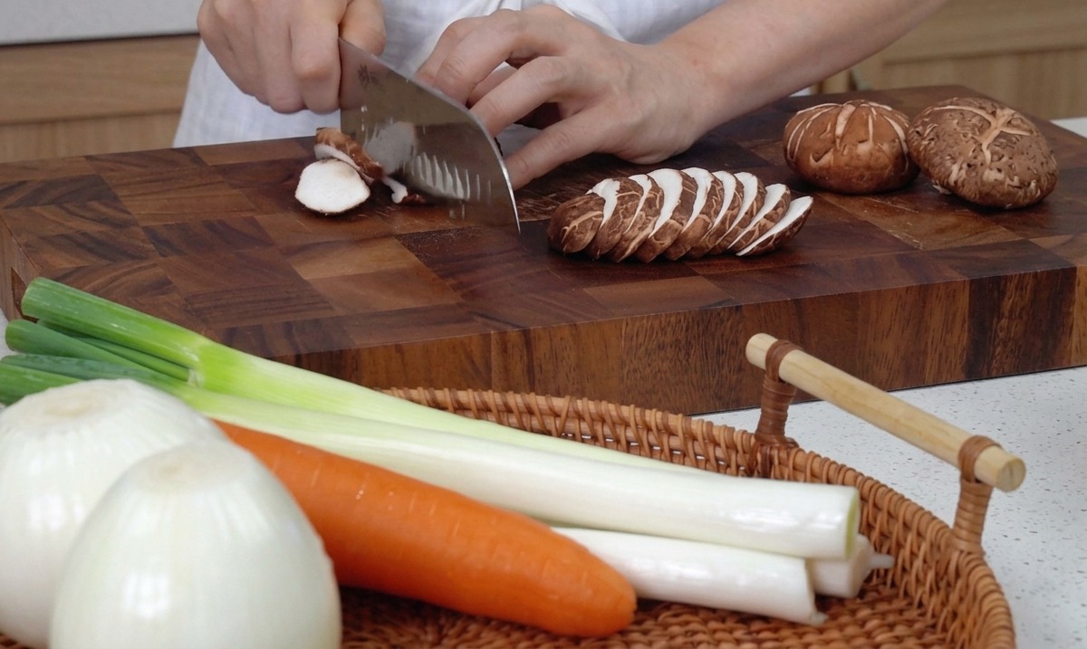 Hände schneiden frische Shiitake-Pilze mit einem großen Küchenmesser auf einem Holzbrett.