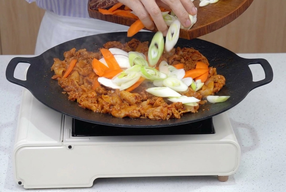 Eine Pfanne mit gebräuntem Schweinebauch, ergänzt um frische Streifen von Karotten, Zwiebeln, Pilzen und Frühlingszwiebeln.