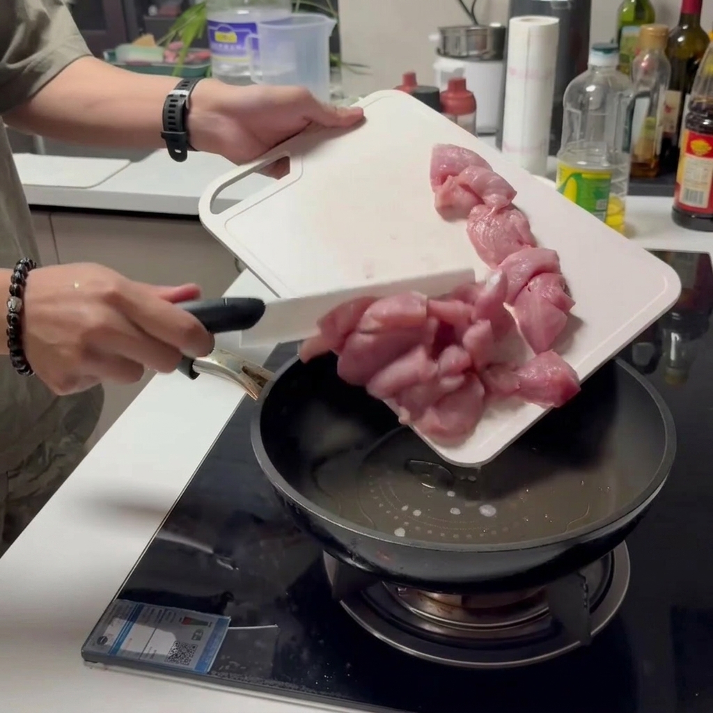 Rohe Schweinefleischstreifen werden von einem weißen Schneidebrett in eine geölte Pfanne geschoben.